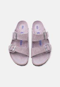 Ζευγάρι ανοιχτό μοβ σουέντ σανδάλια με δύο ρυθμιζόμενες ασημένιες αγκράφες και καμπυλωτές σόλες, σε λευκό φόντο.