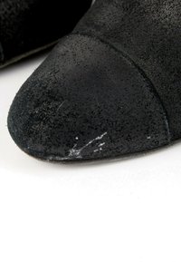 Gros plan sur l'avant d'une chaussure en daim noir avec des marques blanches visibles sur la surface avant, sur fond blanc.
