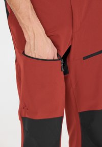 Röda utomhusbyxor med svarta accenter, med en dragkedjeficka. Tillverkade av ett mjukt, flexibelt material, designat för funktionalitet och komfort.