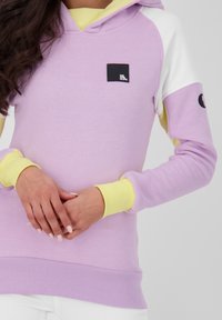 Felpa viola con accenti gialli e bianchi, polsini a coste e una piccola toppa nera con la scritta "WE ARE ALIVE." Tessuto morbido, vestibilità rilassata.
