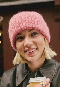 Różowa dziana czapka z prążkowaną teksturą noszona z czarną skórzaną kurtką. Model trzyma kubek z kawą na wynos i ma krótkie blond włosy.