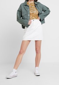 Chaqueta de mezclilla verde, camisa de estampado animal en color marrón dorado, minifalda de algodón blanca y zapatillas Converse blancas con detalles en negro.