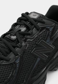 Chaussure de sport noire avec une tige en mesh et des accents brillants, des lacets texturés et un logo bien en évidence. Talon renforcé pour un meilleur soutien.