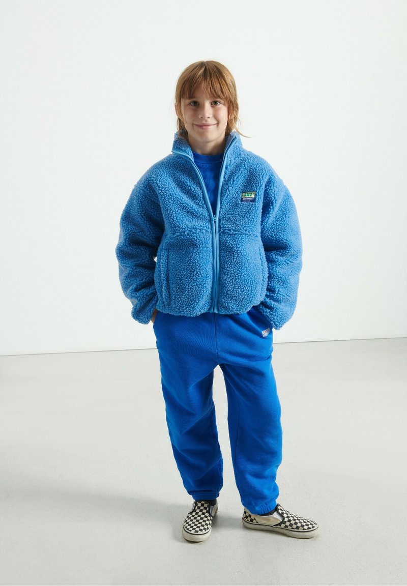 Chaqueta de forro polar azul con material texturizado y esponjoso, cierre de cremallera y logotipo. Combinada con pantalones azules a juego y zapatillas de cuadros sin cordones.