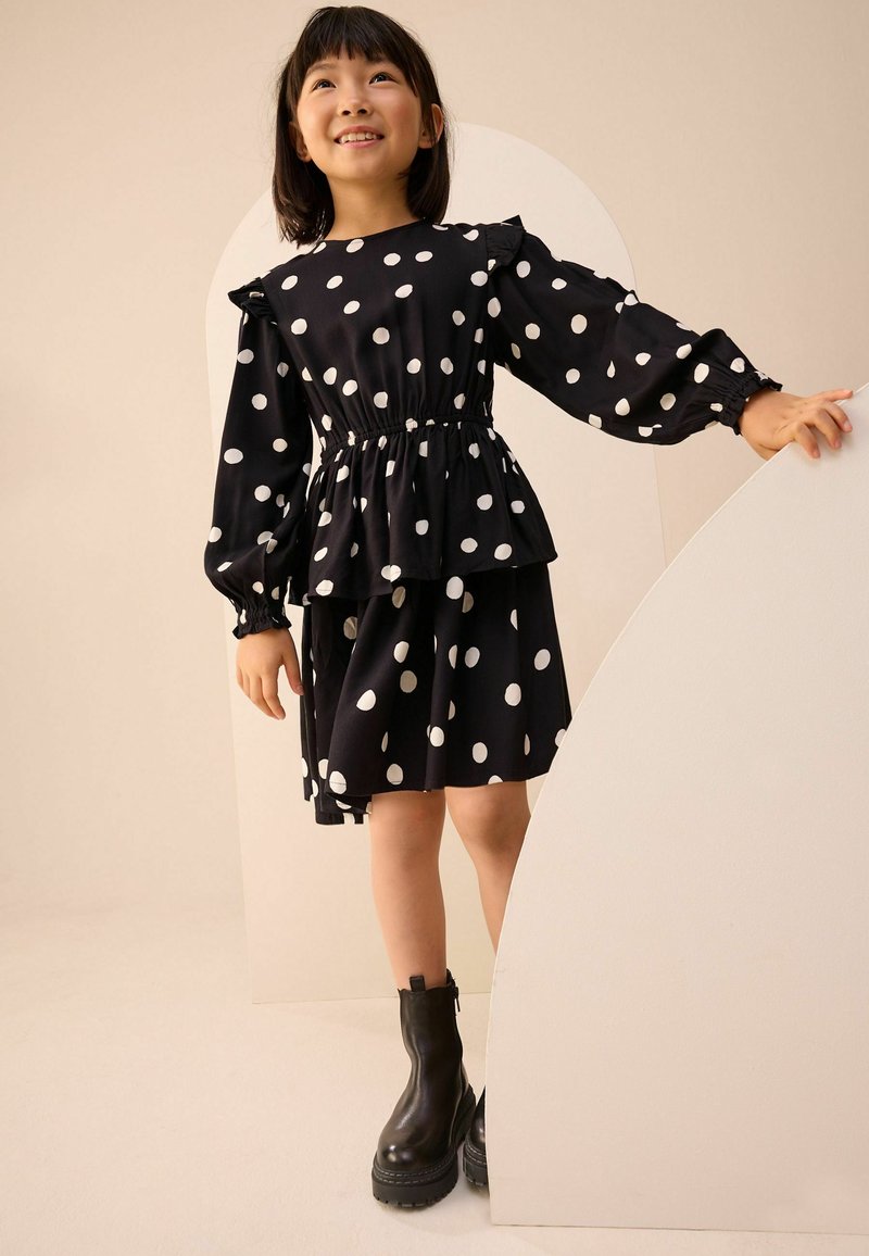 Robe noire à pois avec des manches bouffantes, taille cintrée et ourlet superposé. Associée à des bottines noires à la cheville. Tissu texturé.