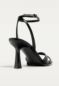 Schwarze High-Heel-Sandalette mit schmalen Riemen und einem Knöchelverschluss. Besitzt ein glänzendes Finish und einen quadratischen, konischen Absatz. Flache Sohle auf der Unterseite.