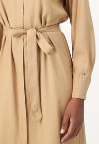 Beżowa sukienka z długim rękawem i ściągniętym w pasie. Posiada pasujący pasek z tego samego materiału, gładką fakturę oraz małe logo na rękawie.