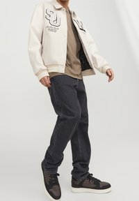 Pánská krémová varsity bunda s černými detaily, šedé tričko, tmavé džíny a černo-hnědé tenisky. Obsahuje výšivku písmen a loga.