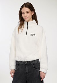 Giovane donna che indossa un pullover bianco in pile con la scritta "New York Athletics Club", abbinato a jeans neri a vita alta, in piedi di fronte a uno sfondo bianco.