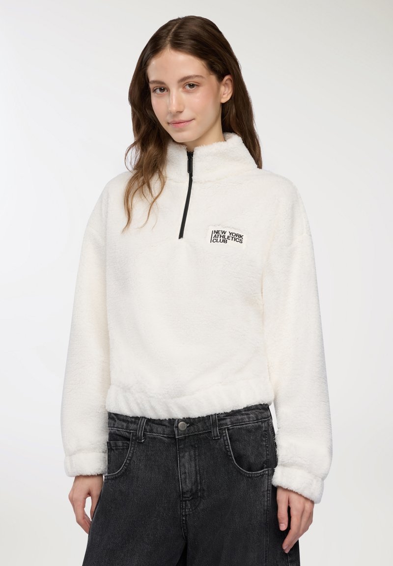 Giovane donna che indossa un pullover bianco in pile con la scritta "New York Athletics Club", abbinato a jeans neri a vita alta, in piedi di fronte a uno sfondo bianco.