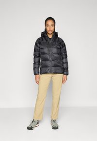 Een vrouw staat met het gezicht naar voren, gekleed in een zwarte gewatteerde jas met capuchon, beige broek en grijze wandelschoenen, tegen een effen grijze achtergrond.