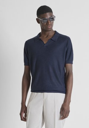 Hombre con camisa tipo polo de punto de manga corta azul oscuro y pantalones plisados beige claro, posando con gafas de sol sobre fondo liso.