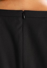 Sort kjole med tekstureret stof, der har en lynlåslukning bagpå og en flad søm langs halsudskæringen. Glat og figursyet silhuet.