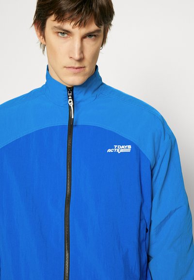 Kék, cipzáras dzseki magas gallérral, könnyű anyagból, kontrasztos vállpanellel és fehér logóval a bal oldalon.