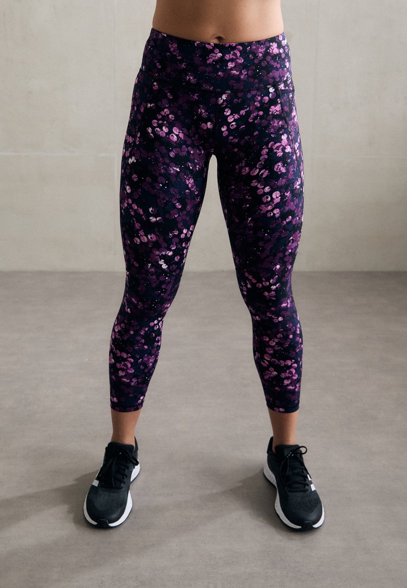 Juodos leggingų su violetiniais ir rožiniais gėlių raštais, turinčių aukštą juosmenį ir priglundantį dizainą, derinamos su juodais sportiniais bateliais.