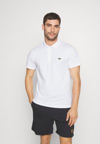 Witte polo met korte mouwen en een kraag met het groene Lacoste krokodil logo. Draagt een zwarte short, met de handen in de zakken.
