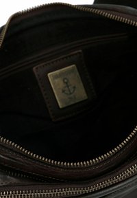 Interno di una borsa in pelle marrone con chiusura a zip, dotata di un'etichetta metallica con design di un'ancora e rivestimento in tessuto scuro.