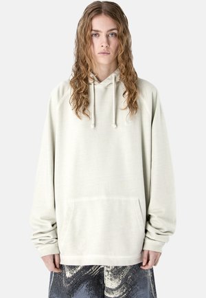 Young person with long curly hair wearing a loose, light beige hoodie with drawstrings and abstract black-and-white patterned pants.