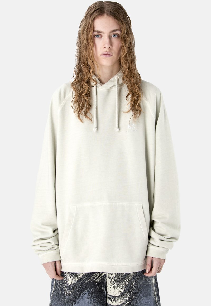 Jeune personne aux longs cheveux bouclés portant un sweat à capuche ample de couleur beige clair avec des cordons et un pantalon à motif abstrait noir et blanc.