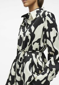 Camisa ceñida de patrón en blanco y negro con cinturón y cuello, que presenta mangas largas y una textura de tela suave. Realzada por detalles de costura.