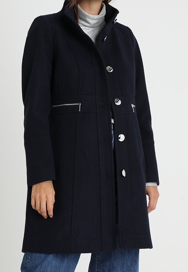 Femme portant un manteau noir longueur genou avec des boutons argentés et des poches zippées sur un col roulé gris et un jean bleu.