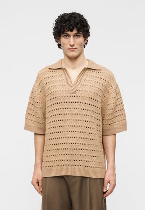 Jeune homme aux cheveux bouclés portant un pull en maille beige à manches courtes avec un motif ajouré et un col large, associé à un pantalon marron ample.