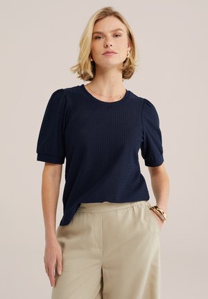 Femme blonde porte un top bleu marine texturé à manches courtes, un pantalon beige à fines rayures, des boucles d'oreilles créoles en or, un collier fin et des bracelets épais, main dans la poche.