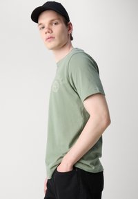 Alyvuogių spalvos medvilninis t-shirt su apvaliu kaklu, trumpomis rankovėmis ir logotipo grafika ant krūtinės. Derinama su juodomis kelnėmis.