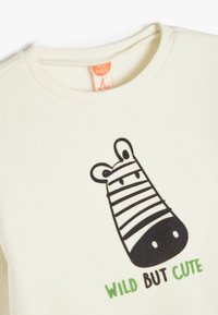 Felpa color crema chiaro con scollo a costine, presenta una grafica di zebra a righe bianche e nere e testo verde che recita "WILD BUT CUTE".