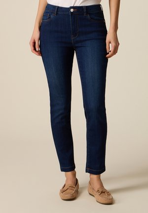 Donna che indossa jeans slim fit blu scuro con tasche frontali e mocassini beige, in piedi su sfondo neutro.