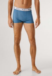 Mann, der barfuß steht und blaue Boxershorts mit einem weißen Bündchen trägt, auf dem "Pepe Jeans" steht, vor einem einfachen hellen Hintergrund.