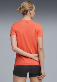 Short-sleeve athletic shirt in bright orange fabric with a smooth texture, featuring a black logo and a relaxed fit. Worn with black shorts.