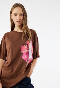 Brun oversized t-shirt med korte ærmer, der har et stort pink og hvidt blomstermotiv på venstre side. Materialet er bomuld, med en afslappet pasform.
