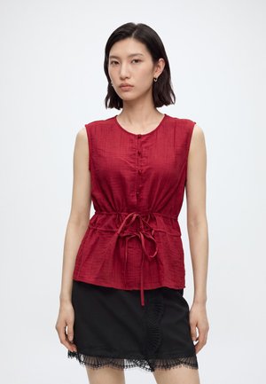 Femme aux cheveux noirs courts portant un chemisier rouge sans manches avec des boutons et une ceinture à nouer, ainsi qu'une jupe noire avec une bordure en dentelle.