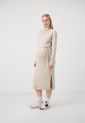 Knit beige maternity dress with long sleeves, side slit, and a tie belt. Worn with white socks and cream sneakers with dark accents.