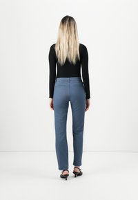 Kvinna med långt blont hår står med ryggen mot oss, klädd i en svart långärmad topp, blå åtsittande byxor och svarta klackskor med kitten-heel.