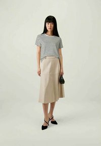 Γυναίκα που φοράει μαύρο και άσπρο ριγέ μπλουζάκι, μπεζ μίντι φούστα, μαύρα μυτερά τακούνια και κρατά μικρή μαύρη τσάντα χειρός.