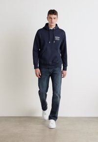 Marineblå hettegenser med lomme foran og snorer, kombinert med mørke jeans og hvite sneakers, på en lys bakgrunn.