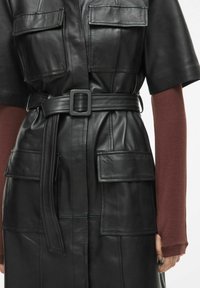 Juodas odinis suknelė su trumpomis rankovėmis, diržu ir keturiomis priekinių kišenėmis. Oda turi lygią tekstūrą ir nežymų blizgesį.