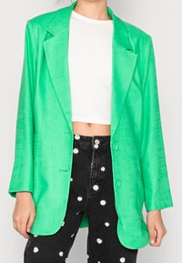 Blazer verde texturizado con solapas de muesca, cierre de un botón y dos bolsillos frontales, combinado con un top corto blanco y jeans negros con flores.