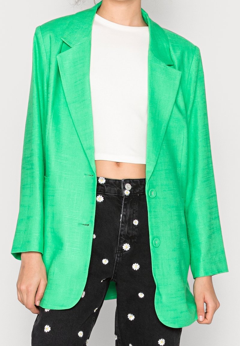 Blazer verde texturizado con solapas de muesca, cierre de un botón y dos bolsillos frontales, combinado con un top corto blanco y jeans negros con flores.