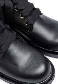 Black leather ankle boots featuring sleek, glossy surface, round toe, and gold eyelets with wide black ribbon laces. Textured fabric accents.