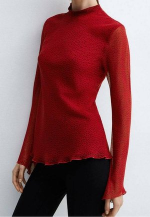 Femme portant un haut rouge transparent ajusté à manches longues avec de petits pois noirs et un col montant, associé à un pantalon noir.
