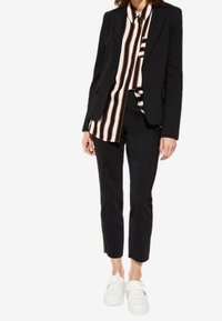 Chaqueta de traje negra ajustada sobre una camisa a rayas marrón y blanca, combinada con pantalones negros hasta el tobillo y zapatillas blancas.
