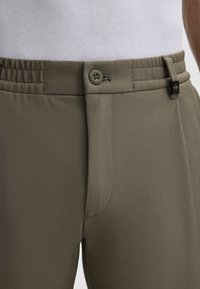 Groene geweven stof broek heeft een aansluitend ontwerp met een enkele knoopsluiting en een zijzak, en toont een gladde, gestructureerde textuur.