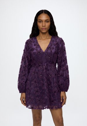 Mujer con cabello largo y oscuro que lleva un mini vestido púrpura transparente con bordados florales, mangas largas abullonadas y escote en V.