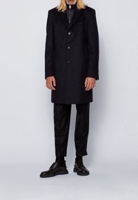 Svart ullkappa med snedslagskrage, enkelknäppt design, två framfickor och fem knappar. Matcherad med svarta byxor och skor.