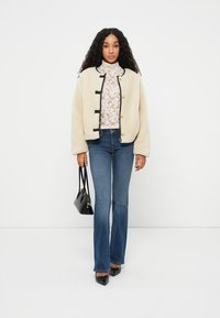 Creme fleecejakke med sort kant, gyldne knapper og en afslappet pasform, parret med mørkeblå flared jeans og en sort håndtaske.