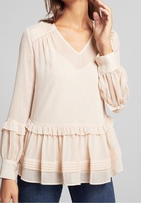 Femme portant un chemisier beige clair à manches longues avec des détails plissés et volantés, associé à un jean bleu foncé.