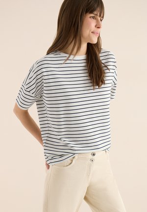 Kvinde iført en hvid t-shirt med korte ærmer og marineblå vandrette striber samt lysebeige bukser, stående med hænderne bag ryggen og kigger til siden.